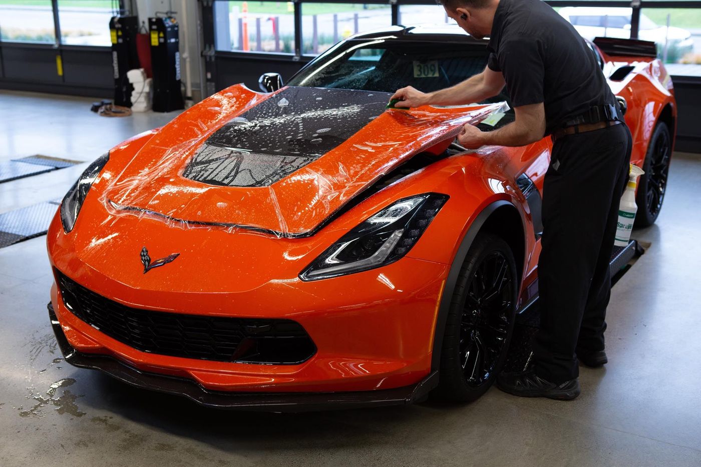 Man applies protective film to a bright orange Corvette hood in a garage.