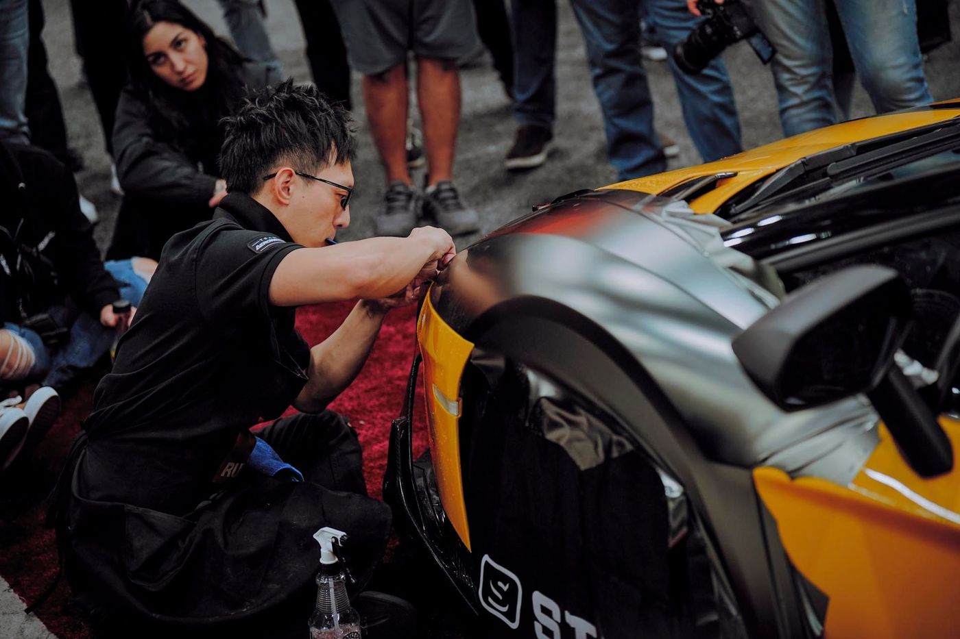 A man applying wrap to a yellow sports car in a crowded setting.